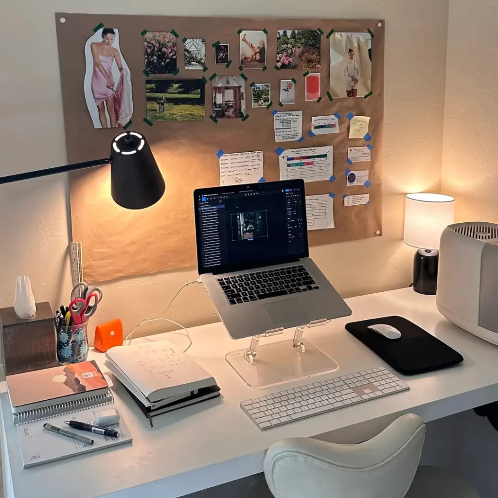 A well-organized workspace featuring a laptop on a clear stand, a keyboard, notebooks, and a bulletin board with pinned inspiration images and notes. A sleek and functional home office setup.